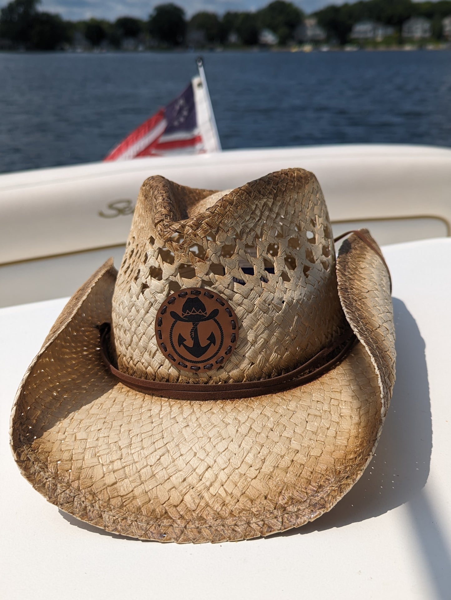 Photo of a Lake Cowboy Signature Cowboy Hat with the iconic hat, lasso and anchor logomark on the front of the hat. The hat is shown sitting on a table on a boat with the American flag in the background. The photo was taken on Lake Minnetonka, MN.